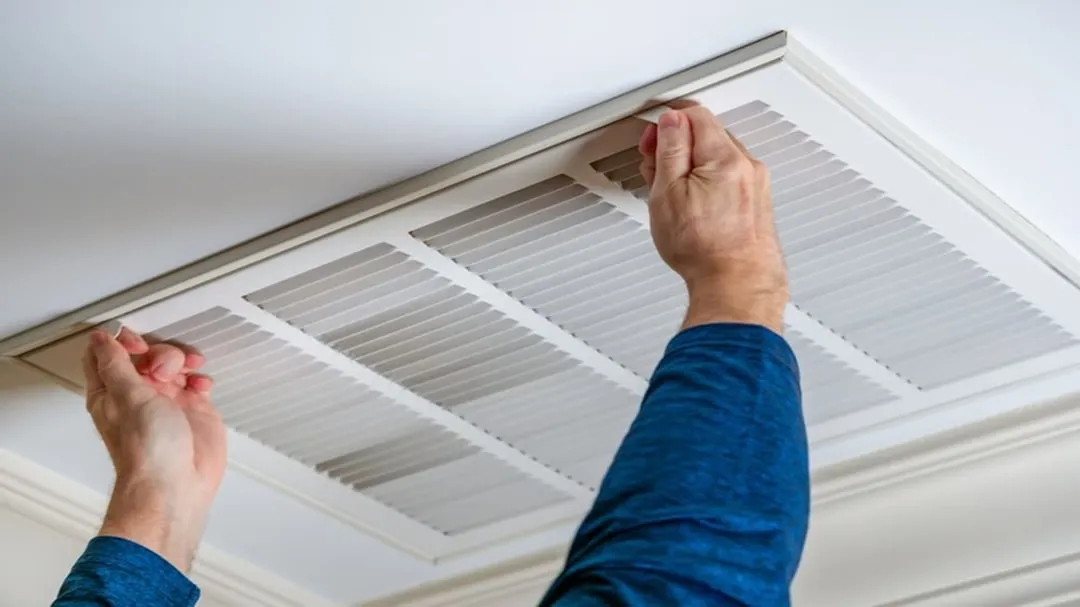 homeowner replacing a pleated air filter in a hallway intake vent - spring ac maintenance to prevent summer failures